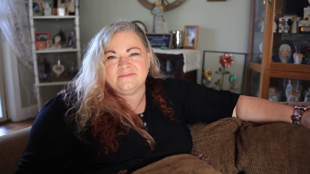 Christina, a smiling white woman with long grey hair looks at the camera. She sits in her living room, with frames and tscoches behind her. She has red dye on the tips of her hair. Her left hand is extended on her couch, with a watch on it.