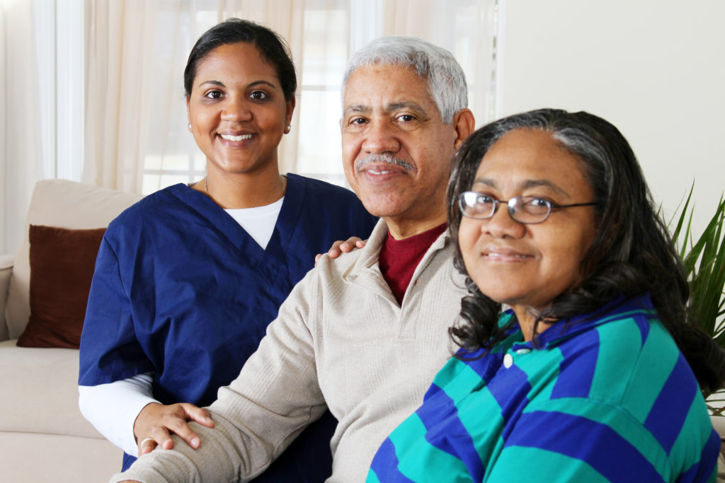 A family of three smiles at the camera. On the left is a female-presenting woman in her 30s with brown skin, wearing navy scrubs. She has her hands supportively on the man to her left, a man in his 70s with grey hair and mustache and brown skin. To his left is a female-presenting person with greying black shoulder-length hair. She appears to be in her 60s, and is wearing a blue and teal striped shirt. Behind them is a plant. A great thing about WA Cares is that is allows family members to get paid for caring for loved ones when they need it.