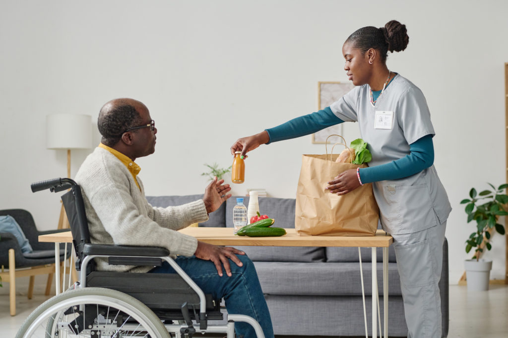 A Black male-presenting person aged around 65 sits in a wheelchair at a kitchen table. He has short, receding hair and glasses. With his left hand he reaches for a bottle that his home care aide is handing him. On the right is a Black female-presenting person aged around 35. She is wearing light blue scrubs with a teal shirt underneath, and has green nail polish. She is unpacking groceries from a brown paper bag on the table. On the table are zucchini, a red pepper, and a bottle of water. They are talking about WA Cares.