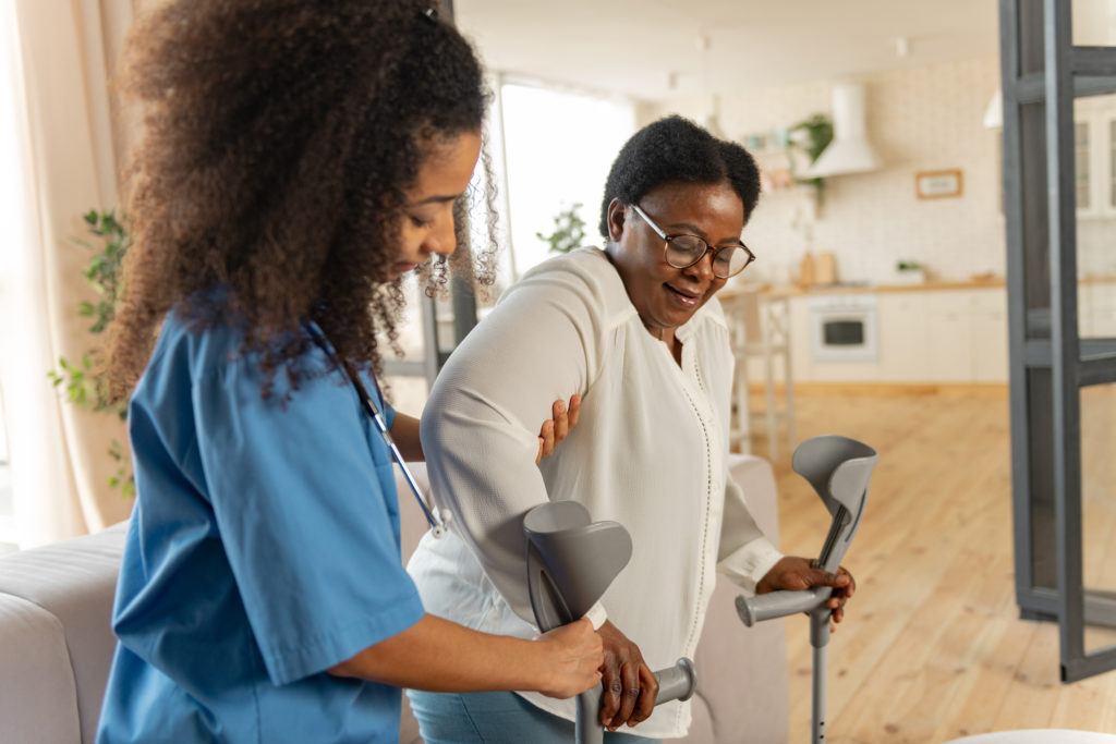 A Black female-presenting woman in her 50s wears glasses and puts her arm into arm braces. To her right, a light skinned female-presenting woman in her 40s supports her right arm, helping her into the brace. She is a medical aide, wearing light blue scrubs with a stethoscope around her neck. In the background is a light airy kitchen.