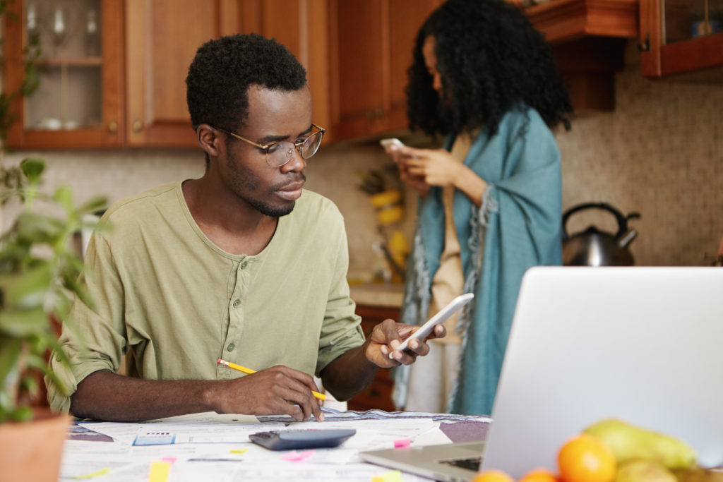 A Black male-presenting person with short hair wears glasses and sits at his laptop at his kitchen table. He has a pencil in his right hand. He is using his phone in his left hand and looking at it. In front of him, around his laptop, are papers and calculaors on the table. Behind him is a Black person with long hair standing against a kitchen counter. There is a tea kettle behind them.  