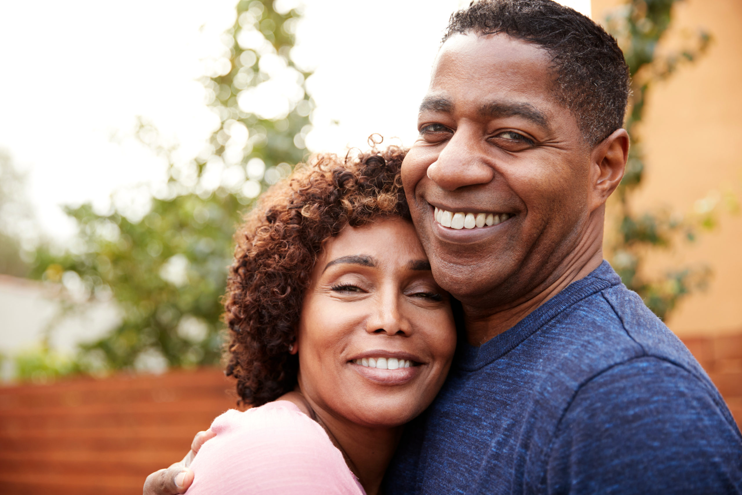 A close up of a smiling Black couple hugging. On the left is a female-presenting person with a pink shirt and curly shoulder-length hair. On the right is a male-presenting person in a blue shirt. They know they'll be covered by the WA Cares Act if they ever need long-term care.