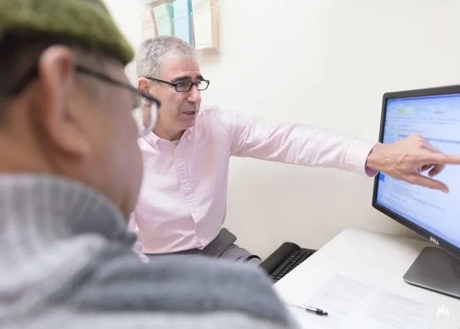 El Dr. Charles Mayer, un hombre de raza blanca de aproximadamente sesenta años, señala la pantalla de una computadora con la mano izquierda. Usa gafas negras y una camisa de color rosa de manga larga. A su izquierda se ve la espalda de un paciente, que usa gafas y un gorro verde. Está mirando la pantalla donde señala el Dr. Mayer, quien le explica los beneficios de WA Cares.