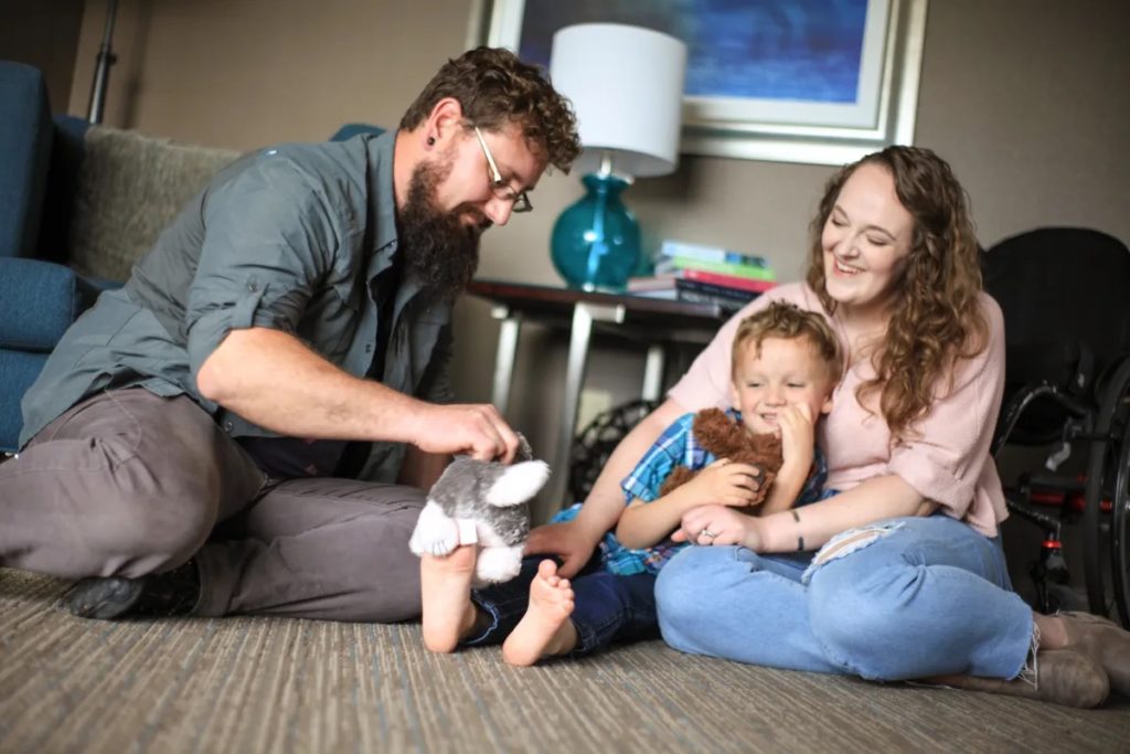 A young family of three sits and plays on their floor. Dani, an advocate for WA Cares, sits on the floor with her legs folded. She is white skin and has long curly brown hair and a pink shirt, and is smiling. She is holding her son in her lap, who in sitting on the floor with his legs out straight. He is a white boy aged around 6. To the left is Dani's husband, a white man with glasses and short brown curly hair and a beard. He is playing with a stuffed animal on his son's foot. Behind them is a table with a blue lamp and a stack of books.