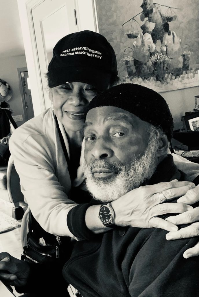 A Black woman in her 60s, Nina, stands behind her husband and hugs him. She is wearing a watch and black hat that says "well behaved women rarely make history" and smiling. Her husband, a Black man in his 60s, is looking at the camera. He has a white beard and is wearing a black skull cap. 