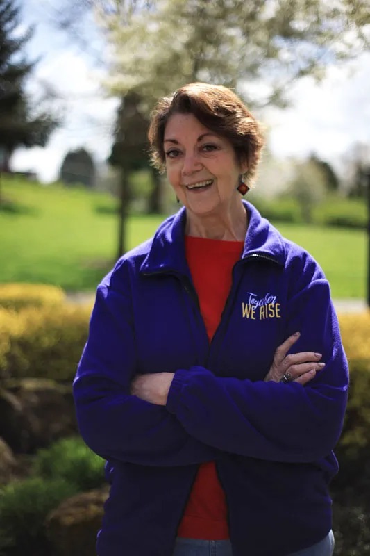 A white woman named Vicki smiles at the camera in a green park. She is in her 60s, with short brown hair. She is wearing a purple fleece that says "We Rise" over a red shirt. Her arms are folded.