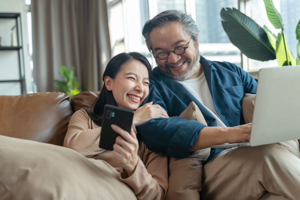 A light skinned female-presenting person in her 30s smiles on a brown couch and holds a smart phone. She is leaning on a light skinned male-presenting man in his 60s, with her right hand on his arm. The man has grey hair and beard and is wearing glasses, and holds a laptop in his lap. He is also smiling. Behind them are plants and a window with daylight coming in.