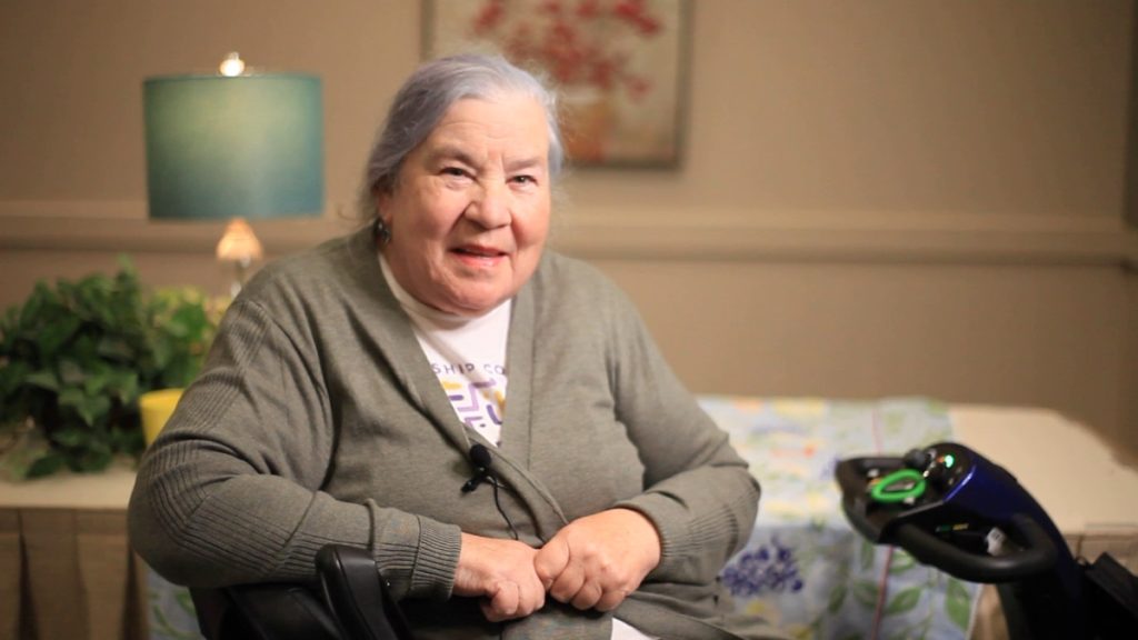 A white woman named Kathy smiles at the camera in her house. She is in her 70s with long grey hair in a pony tail, and sitting on an electronic walker. Behind her is a lamp with a blue shade and a picture of red flowers.