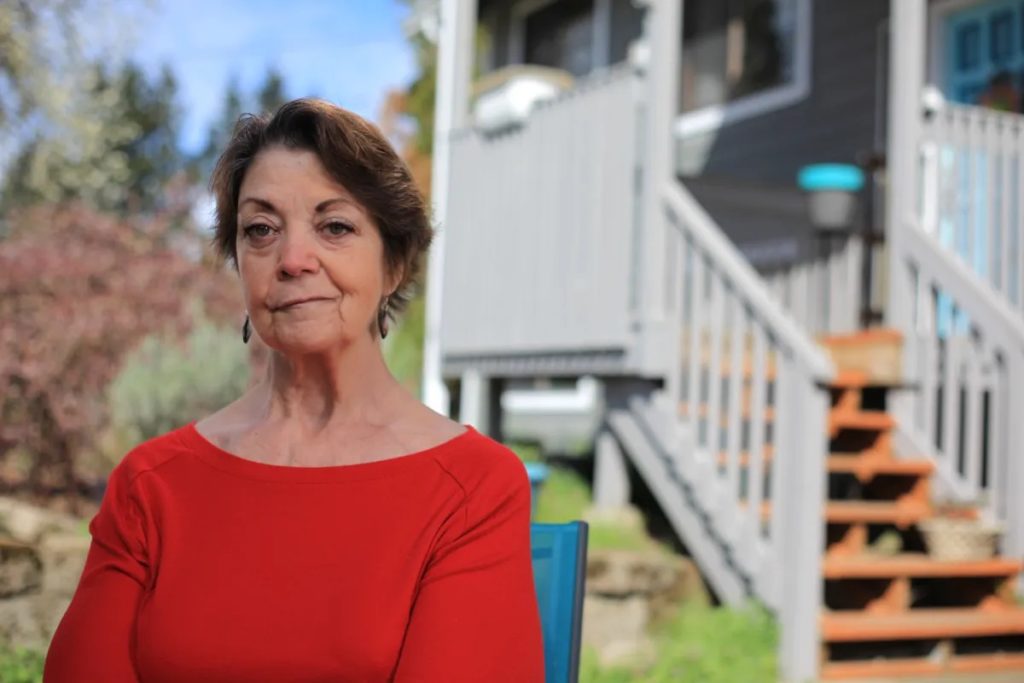 A white woman named Vicki, an advocate for WA Cares, looks with resolve at the camera. She is in her 60s. She has short brown hair and a red shirt. She is standing in her backyard, with stairs up to her porch behind her.