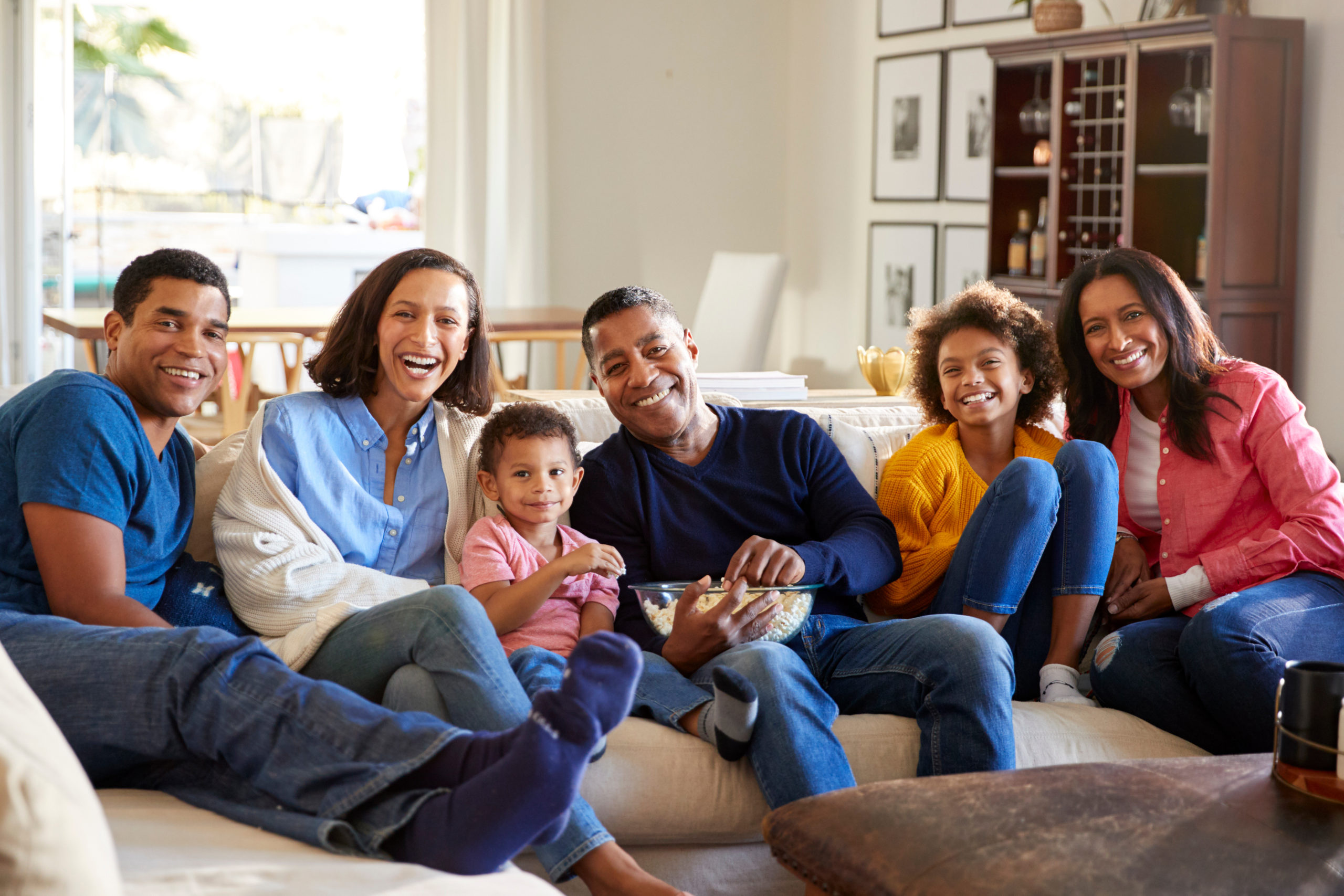 3 generations of supported by WA Cares sit on a couch in their living room. They smile and look at the camera. In the center is a mixed race male-presenting person aged approximately 50. He is smiling and holding a bowl of popcorn. To his right is a child aged around 4, wearing a pink shirt. They are surrounded by four mixed-race family members, 3 female-presenting and one male-presenting.