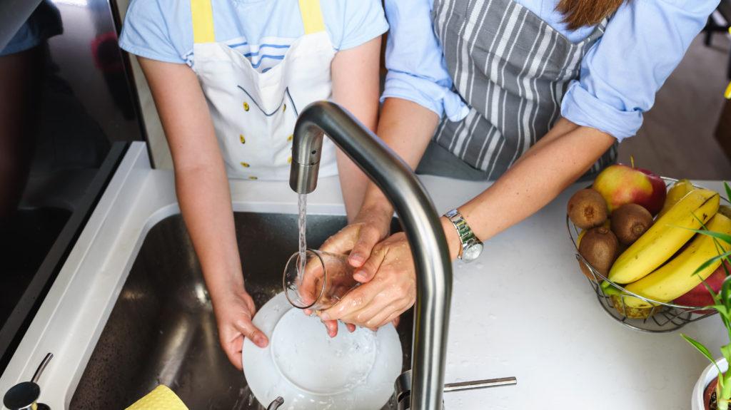 Two people wear aprons and wash dishes together at a kitchen sink. Both of their hands are in the water at the same time.